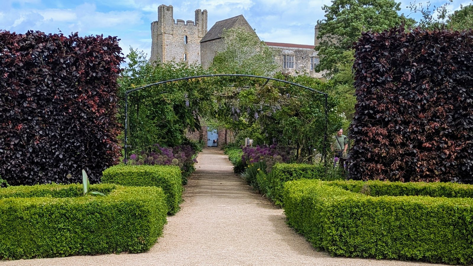 The medieval ruins of Helmsley Castle looks down over the green, lush beauty of Helmsley Walled Garden. Neatly trimmed hedges either side of a gravel path lead towards a plant-covered archway. The castle is behind the wall in the background.